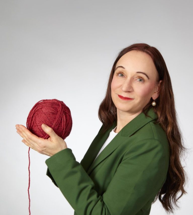 Birgit Cyba mit einem roten Wollknäuel in der Hand, von dem ein Faden nach unten hängt